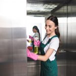 beautiful smiling young janitor cleaning elevator with detergent and rag